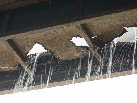Corroded commercial box gutter leaking heavily during rain, with rusted holes and failing waterproofing